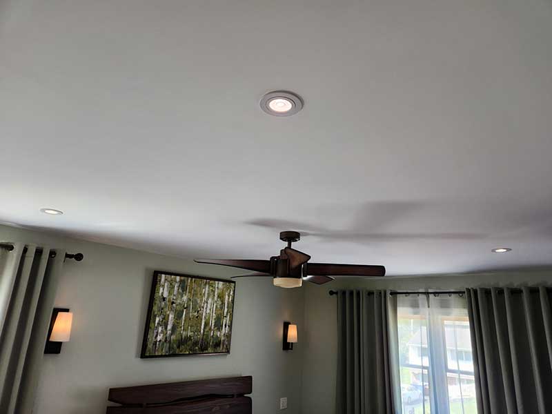 Ceiling fan installation in a modern room with recessed lighting, green curtains, and a wall-mounted artwork featuring trees.