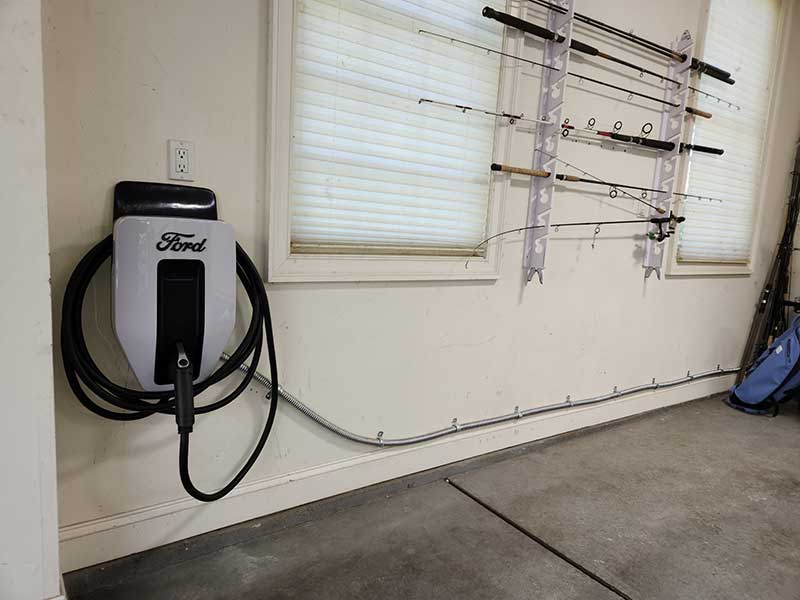 EV charger mounted on a wall, featuring a Ford logo, with fishing rods displayed in the background, illustrating residential EV charger installation services by Nalset Electrical Services.