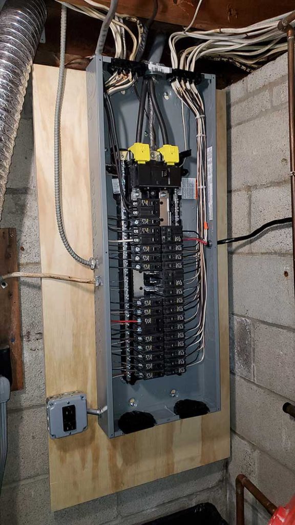 Main electrical panel installation with multiple circuit breakers, wires neatly organized, mounted on a wooden backing in a basement setting.